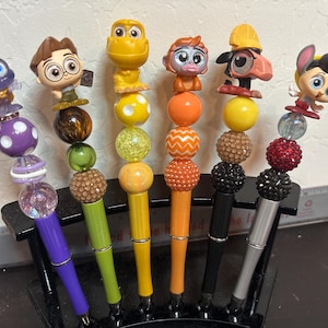 Puede incluir: Una colección de seis bolígrafos decorativos con adornos de personajes. Cada bolígrafo presenta un diseño único con cuentas de colores y una figura de personaje. Los bolígrafos se exhiben en un portalápices negro.