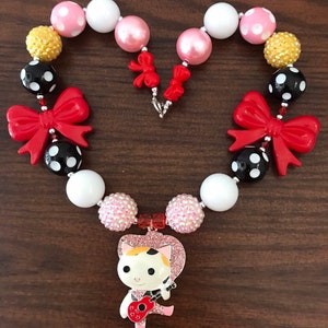 May include: A heart-shaped necklace with a pendant of a cartoon cat playing a red guitar. The necklace features a variety of beads in pink, white, black with white polka dots, and gold. Red bows accent the design.