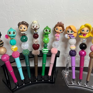 May include: Ten colorful pens with different Disney character toppers. Each pen has a variety of beads and charms attached to it. The pens are displayed on a black and clear stand.