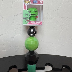 May include: A green and black decorative stand featuring a Bitty Pop! figure of Oogie Boogie from The Nightmare Before Christmas. The stand has a series of black and green spherical beads and a green cylindrical base. The figure is in its original packaging.