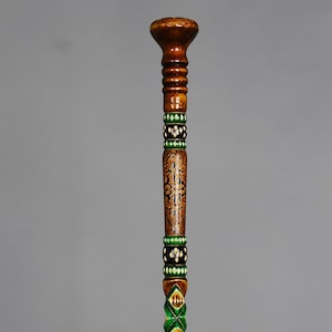 Może przedstawiać: Drewniana laska z ozdobnymi rzeźbami i zielonymi i czarnymi akcentami. Górna część laski jest zaokrąglona, a dolna część jest rzeźbiona tak, aby przypominała węża.