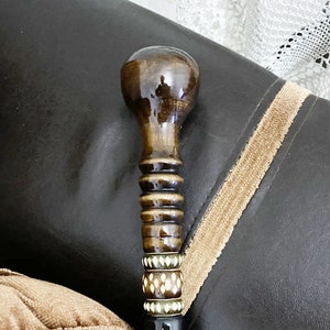 Op de afbeelding: Een houten wandelstok met een bruin, afgerond uiteinde en ingewikkelde inlegwerkontwerpen in goud en wit. De stok is zwart met een goud en wit patroon.