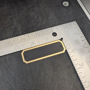 a blank plate ready to be engraved shown in a photograph with a ruler to confirm the size is 2.75 in by 875 in this item has a gold border with a black background along with two holes for mounting and gold screws