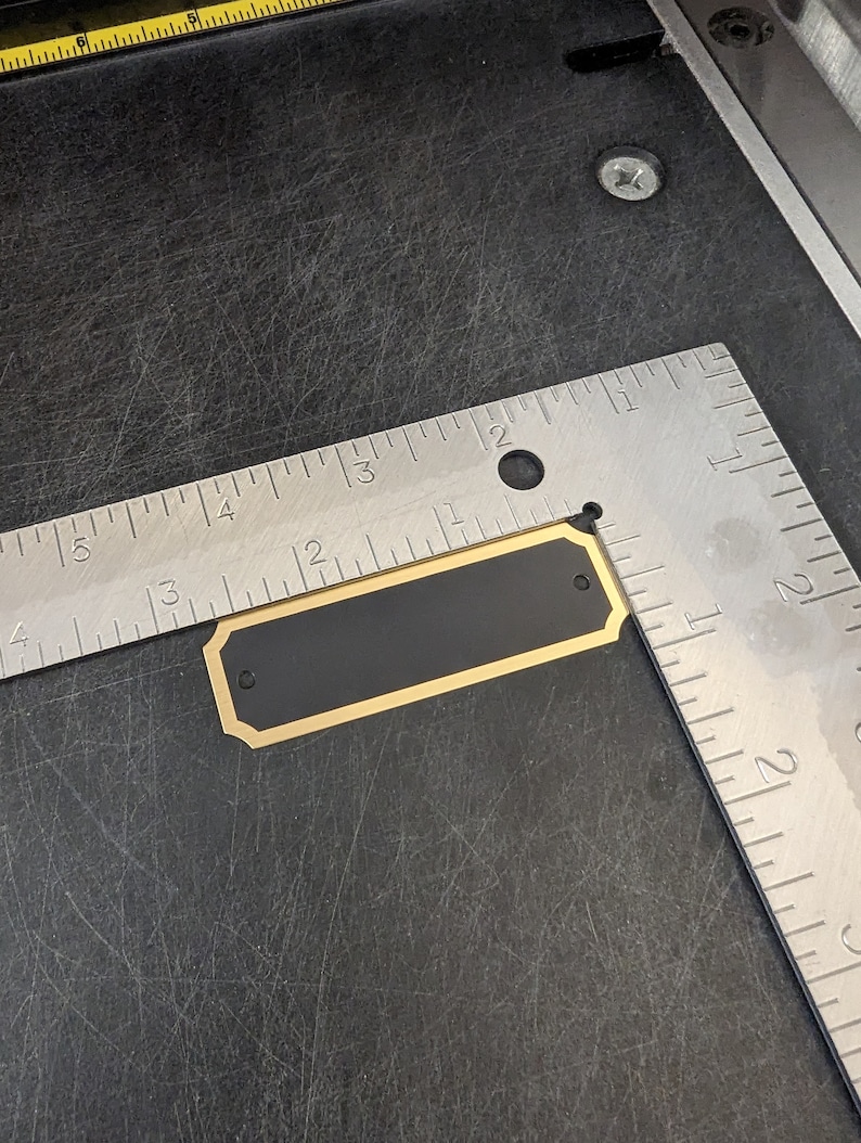 a blank plate ready to be engraved shown in a photograph with a ruler to confirm the size is 2.75 in by 875 in this item has a gold border with a black background along with two holes for mounting and gold screws