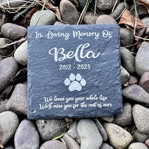 May include: A square dark gray slate memorial plaque featuring the engraved text In Loving Memory Of Bella 2012 - 2025, a paw print icon, and the phrase We loved you your whole life We'll miss you for the rest of ours in white lettering.