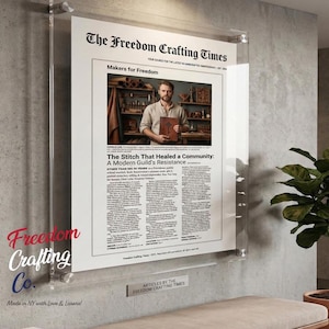 May include: A framed newspaper clipping titled "The Freedom Crafting Times" with the headline "The Stitch That Healed a Community: A Modern Guild's Resistance." The image features a man holding a book. The frame is mounted on a gray wall.
