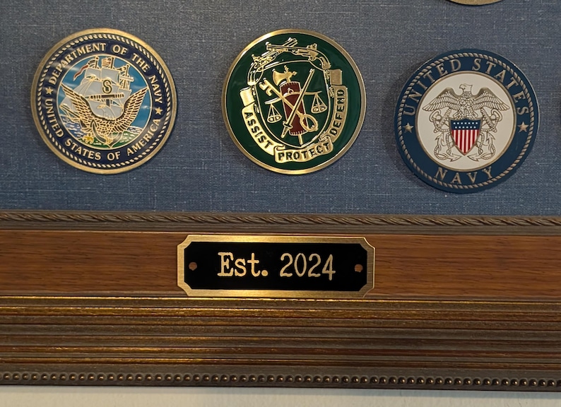 an example of a brass engraved plate being used on a picture frame to notate when something was established the item is a black rectangular plate with a gold border and gold text it also features two holes for mounting with small gold screws