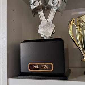 a black brass engraved tag shown in use on a trophy the tag is rectangular in shape with slightly notched corners a gold border and two holes for mounting