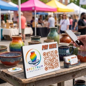 May include: A white rectangular sign with the text Pay Here and a Spectrum Pottery logo featuring a rainbow-colored vase on a wheel, alongside a QR code. The sign sits on a wooden table surrounded by various ceramic bowls, mugs, and business cards.