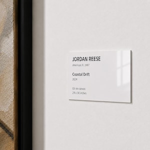 May include: A rectangular white museum wall label with black text reading JORDAN REESE, American, b. 1987, Coastal Drift, 2024, Oil on canvas, 24 x 30 inches. The label is mounted next to a framed painting with a black border and tan tones.