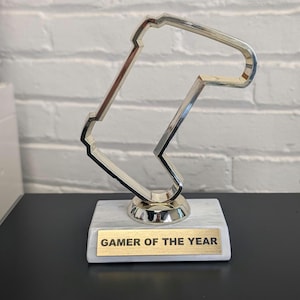 May include: A silver trophy with a unique, angular design, mounted on a white marble base. The trophy has a gold plaque that reads "GAMER OF THE YEAR". The trophy is displayed on a black surface against a white brick wall.