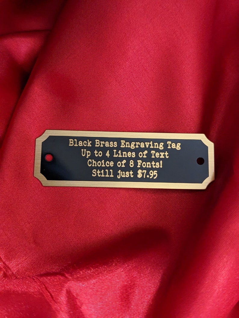 an engraved brass plate featuring a gold brass border and black background with gold text the item is a rectangular shape with slightly notched corners it also depicts two holes used with screws for mounting the item is on a red satin background