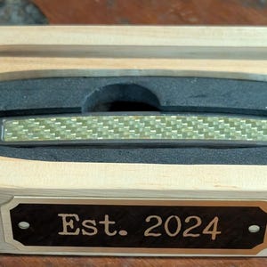 a black brass engraving plate being shown with some custom text so that people can see it can be used to personalize a gift in this case a wooden case for a small pocket knife