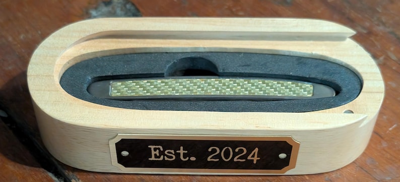 a black brass engraving plate being shown with some custom text so that people can see it can be used to personalize a gift in this case a wooden case for a small pocket knife