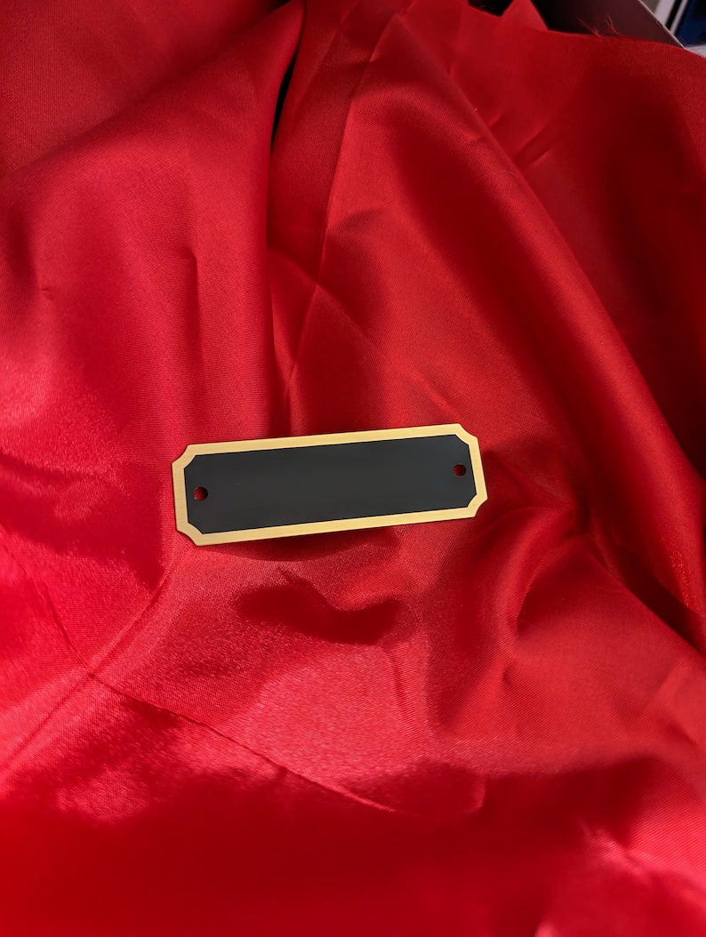a blank engraving tag rectangular and shape with slightly notched corners and two holes for mounting with small gold screws shown on a red satin background