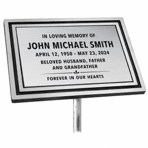 May include: A rectangular silver-toned metal memorial plaque with a black border and a central stake. The text reads: IN LOVING MEMORY OF JOHN MICHAEL SMITH, APRIL 12, 1950 – MAY 23, 2024, BELOVED HUSBAND, FATHER AND GRANDFATHER, FOREVER IN OUR HEARTS.