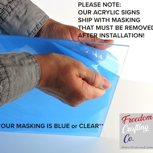 Op de afbeelding: Een doorzichtig rechthoekig acrylbord met een blauwe beschermfolie die gedeeltelijk is losgetrokken. De tekst op de afbeelding luidt: PLEASE NOTE: OUR ACRYLIC SIGNS SHIP WITH MASKING THAT MUST BE REMOVED AFTER INSTALLATION! en **OUR MASKING IS BLUE or CLEAR**