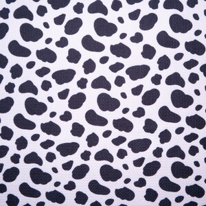 May include: Close-up of a white fabric with a repeating pattern of black, irregular shapes resembling animal spots. The pattern is evenly distributed across the surface, creating a high-contrast, textured design. The fabric appears to be tightly woven.