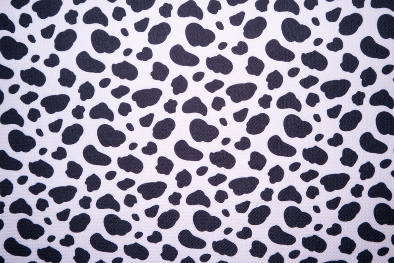 May include: Close-up of a white fabric with a repeating pattern of black, irregular shapes resembling animal spots. The pattern is evenly distributed across the surface, creating a high-contrast, textured design. The fabric appears to be tightly woven.