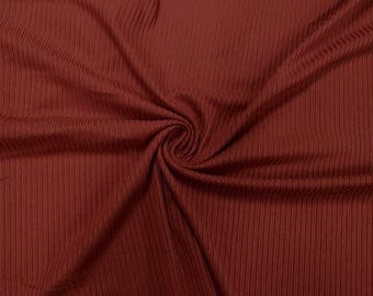 May include: Close-up of a rust-colored ribbed knit fabric. The fabric is folded and swirled, creating a textured pattern. The vertical ribs run the length of the fabric, adding visual interest. The material appears soft and drapes naturally.