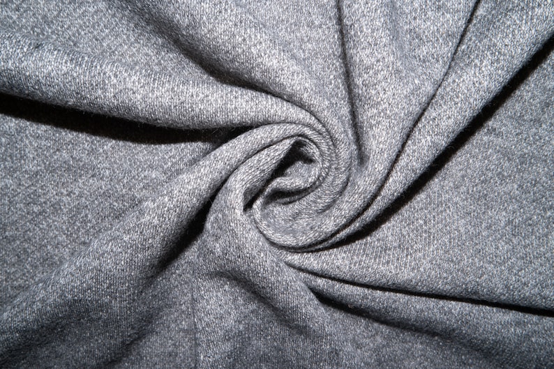 Op de afbeelding: Close-up van een grijze, textuurrijke stof met een draaiende structuur. De stof is zacht en heeft een licht pluche textuur.