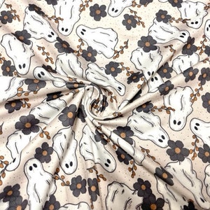 May include: Cream-colored fabric with a repeating pattern of white cartoon ghosts, dark gray flowers, and brown sprigs. The ghosts have simple, oval eyes and a friendly appearance. The fabric has a soft, slightly wrinkled texture.