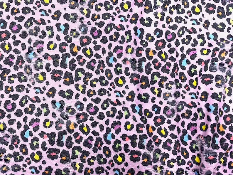 May include: Close-up of a pink fabric with a leopard print design. The black spots are filled with various colors, including yellow, orange, blue, and purple. The fabric has a distressed, torn appearance.