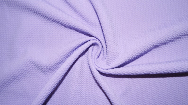 May include: Close-up of a swirl of light purple fabric with a textured, dimpled surface. The fabric is tightly swirled, creating a pattern of folds and shadows. The material appears soft and pliable, suitable for clothing or crafts.