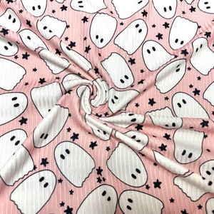 May include: Pink fabric with a repeating pattern of white cartoon ghosts and black stars. The ghosts have simple, oval eyes. The fabric has a ribbed texture, and the pattern is evenly distributed across the surface.