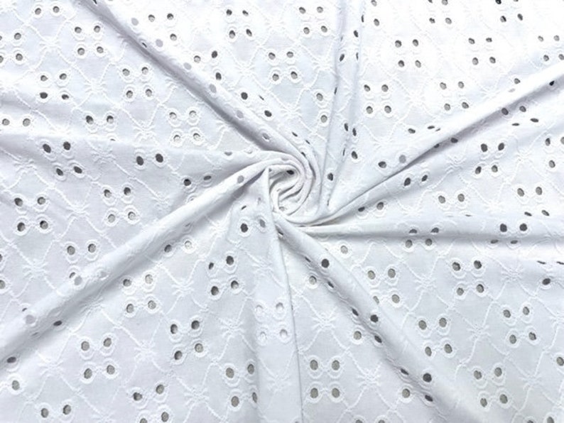 May include: White eyelet fabric with a repeating floral and dot pattern. The fabric is folded and draped, showing the texture and design. The eyelet holes are evenly spaced throughout the fabric.