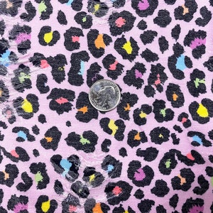 May include: Close-up of a pink fabric with a black leopard print design. The leopard spots are filled with various colors, including yellow, orange, blue, and purple. A quarter coin is placed on the fabric for size reference. The fabric has some distressed areas.