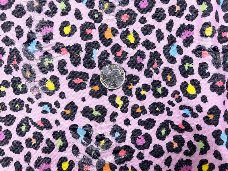 May include: Close-up of a pink fabric with a black leopard print design. The leopard spots are filled with various colors, including yellow, orange, blue, and purple. A quarter coin is placed on the fabric for size reference. The fabric has some distressed areas.