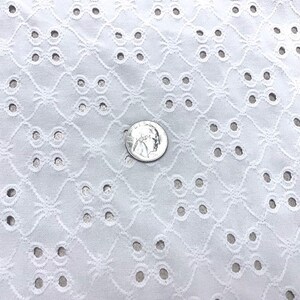 May include: Close-up of white eyelet fabric with a repeating geometric pattern of circles and floral shapes. A silver coin is placed on the fabric for scale, showing the intricate details of the textile. The fabric is likely used for clothing or home decor.