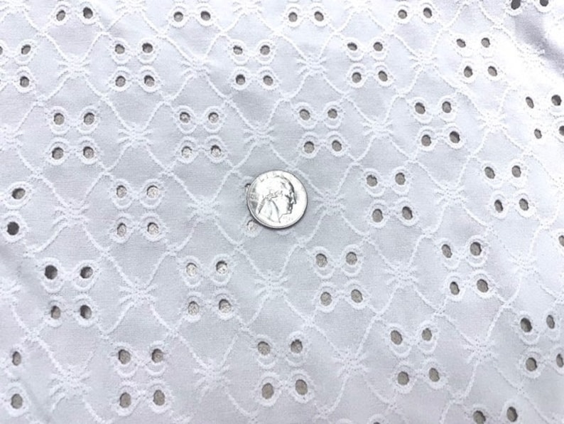 May include: Close-up of white eyelet fabric with a repeating geometric pattern of circles and floral shapes. A silver coin is placed on the fabric for scale, showing the intricate details of the textile. The fabric is likely used for clothing or home decor.