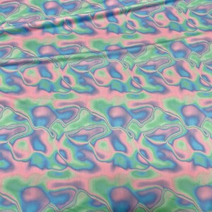 May include: Fabric with a wavy, abstract pattern in pastel colours. The design features flowing shapes in shades of pink, blue, and green, creating a soft, iridescent effect. The fabric appears to be lightweight and flowing.