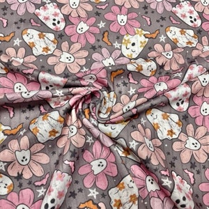 May include: A fabric with a repeating pattern of pink flowers, smiling ghosts, bats, and stars on a gray background. The flowers have a light pink center and darker pink petals. The ghosts are white with orange accents. The fabric has a slight sheen.