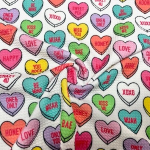 May include: White fabric covered in colorful candy hearts with various phrases like "Kiss Me," "Be Mine," and "XOXO." The hearts are in shades of pink, green, yellow, and purple, creating a playful and romantic pattern.