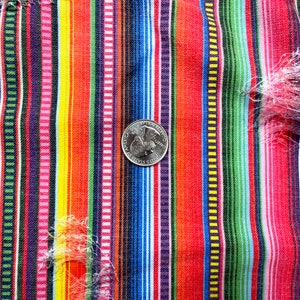 K&ouml;nnte beinhalten: Nahaufnahme eines farbenfrohen Stoffes mit Streifen und einer M&uuml;nze als Ma&szlig;stab. Der Stoff weist vertikale Streifen in Rot, Rosa, Gelb, Gr&uuml;n, Blau und Lila auf. Der Stoff hat einen Used-Look mit ausgefransten Kanten.