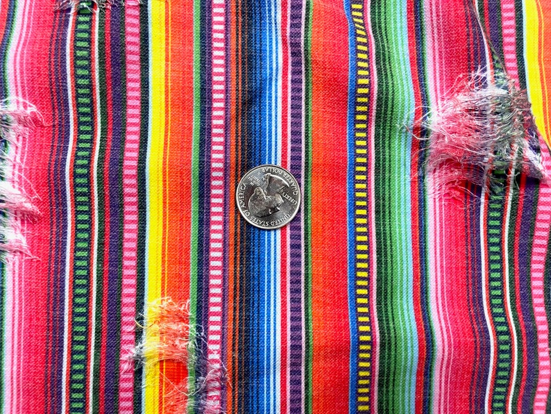 K&ouml;nnte beinhalten: Nahaufnahme eines farbenfrohen Stoffes mit Streifen und einer M&uuml;nze als Ma&szlig;stab. Der Stoff weist vertikale Streifen in Rot, Rosa, Gelb, Gr&uuml;n, Blau und Lila auf. Der Stoff hat einen Used-Look mit ausgefransten Kanten.