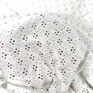May include: Close-up of white eyelet fabric with a delicate floral pattern. The fabric is layered, showing the intricate cut-out design and a hint of red fabric underneath. The eyelet fabric is suitable for clothing and craft projects.