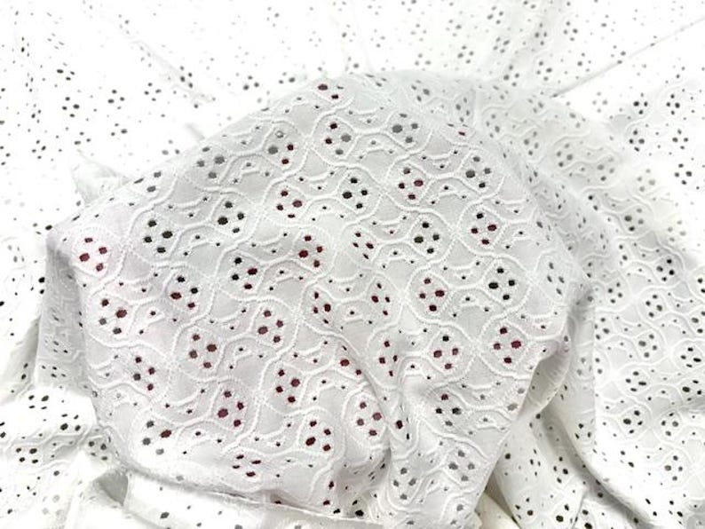 May include: Close-up of white eyelet fabric with a delicate floral pattern. The fabric is layered, showing the intricate cut-out design and a hint of red fabric underneath. The eyelet fabric is suitable for clothing and craft projects.