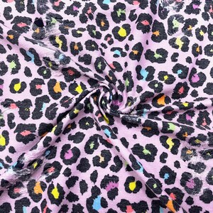 May include: Pink fabric with a black leopard print and colorful accents. The fabric has a distressed, frayed edge. The leopard spots include yellow, orange, blue, and green. This fabric is suitable for apparel and craft projects.