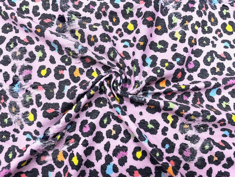 May include: Pink fabric with a black leopard print and colorful accents. The fabric has a distressed, frayed edge. The leopard spots include yellow, orange, blue, and green. This fabric is suitable for apparel and craft projects.
