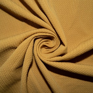 Puede incluir: Primer plano de una tela marrón dorada con un patrón texturizado en forma de gofre. La tela está arremolinada, creando un efecto de espiral. El material parece suave y flexible, adecuado para ropa o decoración del hogar.