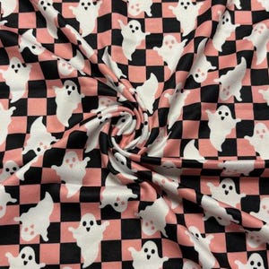 May include: A fabric featuring a repeating pattern of white ghosts with simple black eyes and mouths, set against a checkered background of alternating black and soft pink squares. The material is gathered in the center, creating folds.