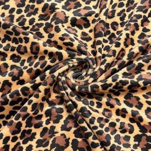 May include: A soft fabric featuring a repeating leopard print pattern in shades of tan, brown, and black. The material is gathered in the center, creating a spiral effect that highlights the texture and the dense, spotted animal print design.