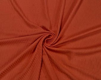 May include: Close-up of a rust-colored fabric with a ribbed texture. The material is gathered in a spiral, creating folds and shadows that highlight the fabric's texture. The color is a warm, earthy tone.