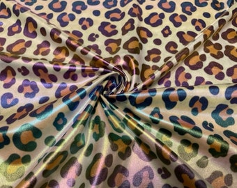 May include: A close-up of a shiny fabric with a leopard print pattern. The fabric has a cream-colored background with brown, purple, and teal spots. The fabric is folded and has a metallic sheen, creating highlights and shadows.