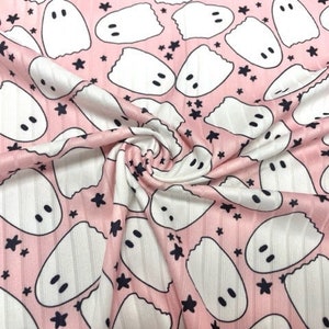 May include: Pink fabric with a repeating pattern of white ghosts and navy blue stars. The ghosts have two black dot eyes. The fabric has a ribbed texture.