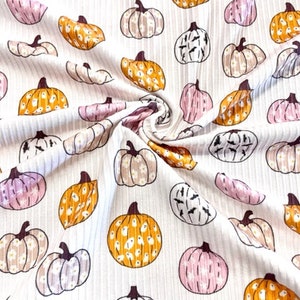 May include: White fabric with a repeating pattern of pumpkins in various colors, including orange, pink, and cream. The pumpkins have different designs and are set against a ribbed background. The fabric is suitable for fall-themed crafts.